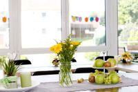 Freundlicher Essbereich; im Vordergrund ist Tischdekoration, gelbe Blumen und eine Obstplatte. Im Hintergrund eine helle Fensterfront.