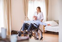 Zu sehen ist ein heller Raum mit einem Bett im Hintergrund. Davor eine Pflegefachkraft, die einen Senioren im Rollstuhl leicht nach hinten kippt. Er hat ausgebreitete Arme; beide lächeln sich an.