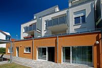 Wohngemeinschaft W&ouml;rt: Ein heller, freundlicher Neubau in wei&szlig; und orange vor blauem Himmel. An zwei Fenstern stehen Blumen. Im Vordergrund befindet sich eine gepflasterte Terrasse.